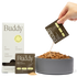 A hand pouring the Buddy daily meal topper powder from a pre-portioned packet into a bowl of kibble dog food, with a box of the product in the background.